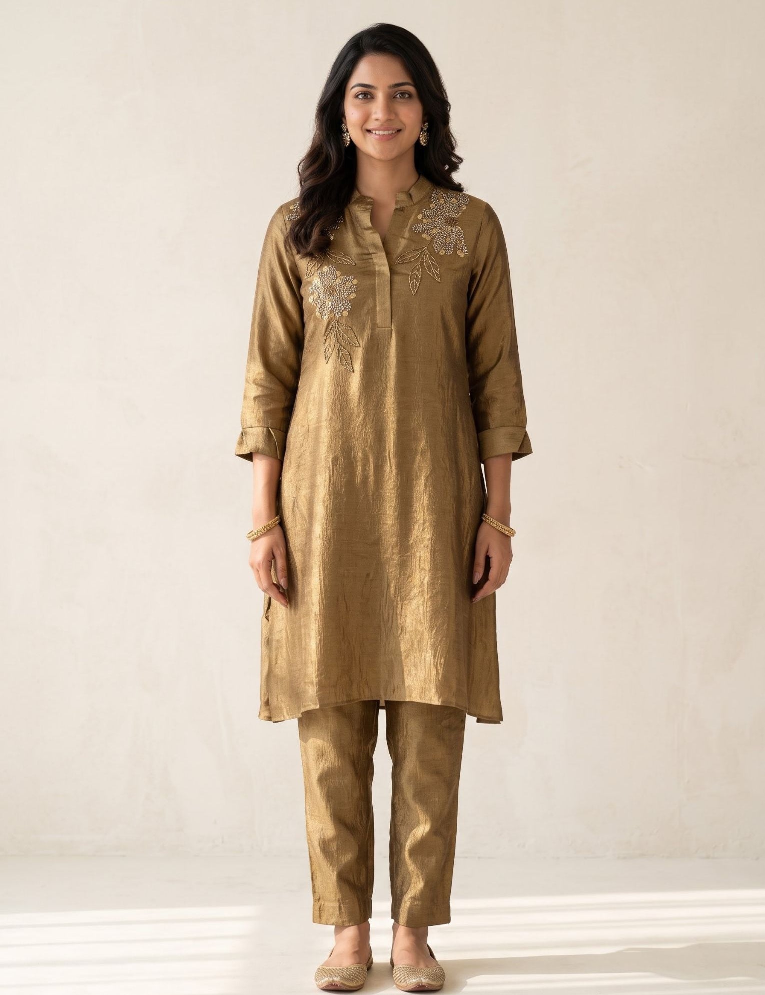 A woman modeling a gold-colored kurta and trouser set with silver floral embroidery on the chest, standing against a neutral beige wall.