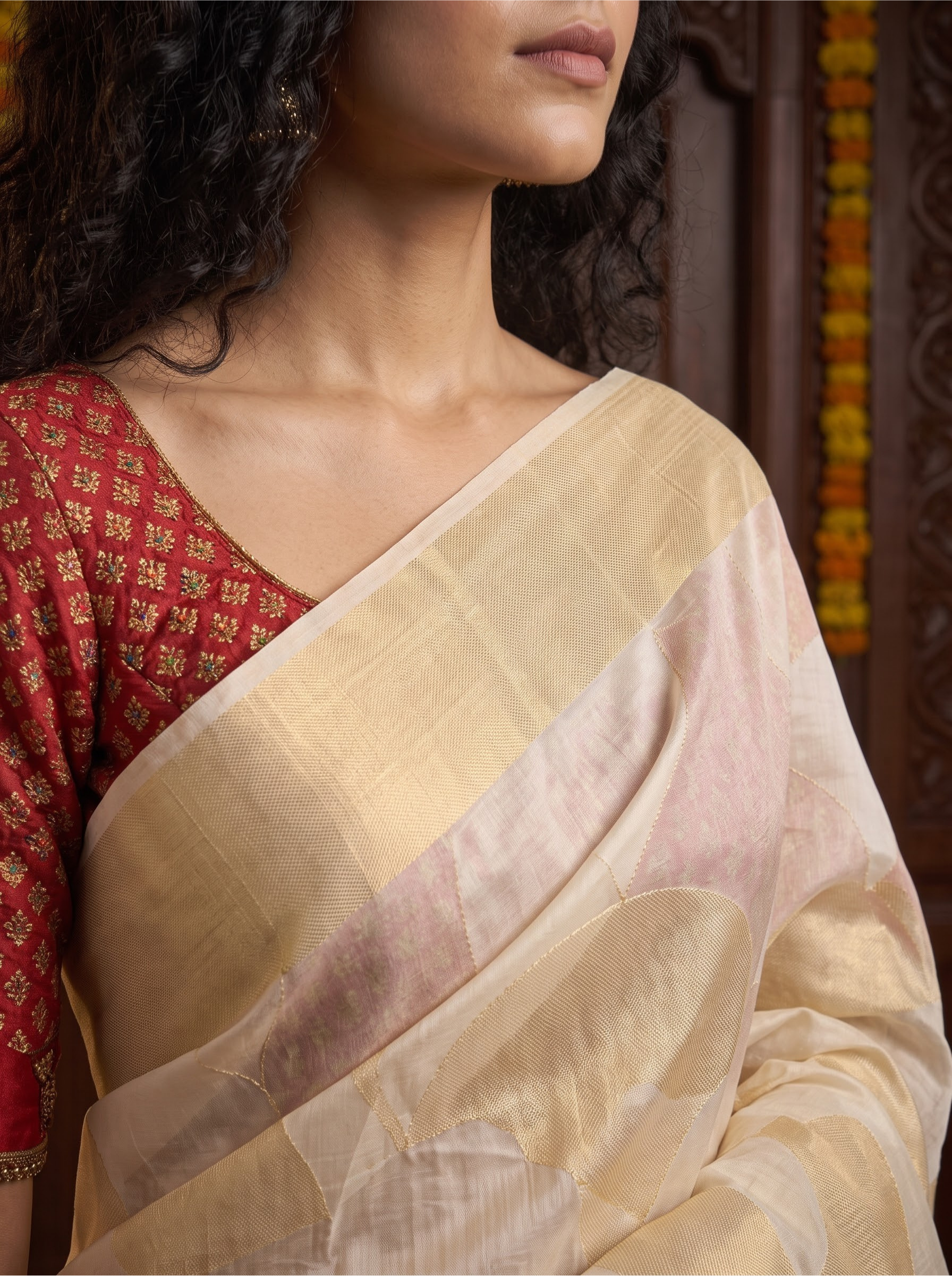A close-up of a woman wearing a soft beige and pale pink saree with a red patterned blouse, set against a dark wooden background.