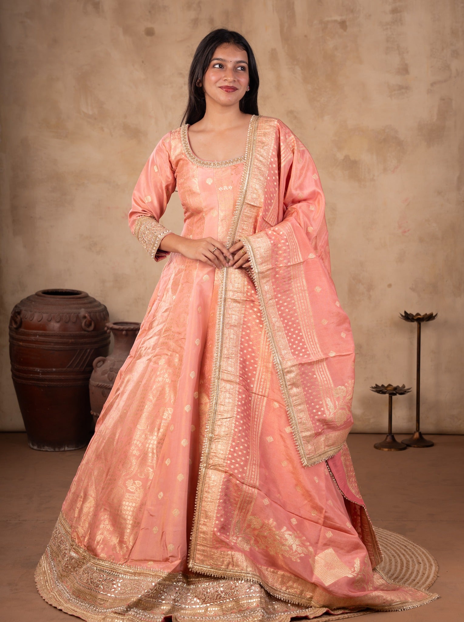 Front view of a model in a blush pink raw silk Anarkali with gold motifs, a wide gold hem and a matching bordered dupatta over one shoulder