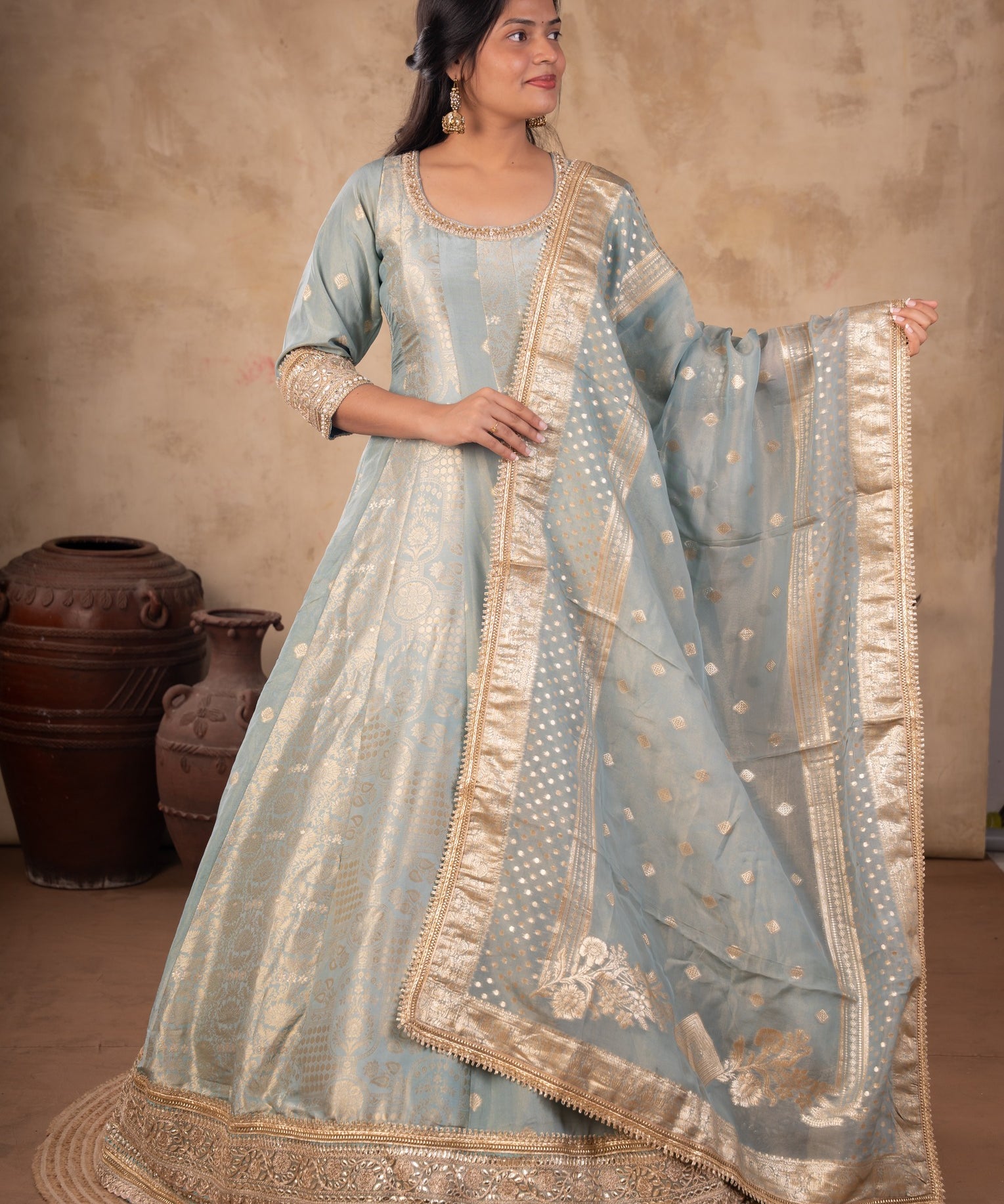 Model wearing a pale blue raw silk Anarkali with gold motifs, an ornate gold hem, and a matching dotted dupatta with a wide gold border.