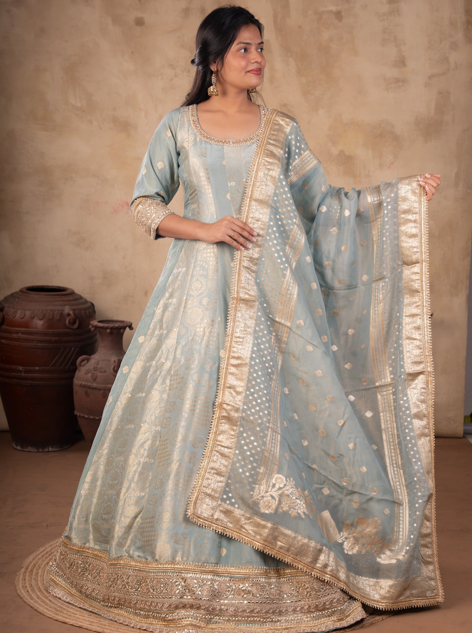 Front view of a model in a pale blue raw silk Anarkali with gold motifs, an ornate gold hem and a dotted dupatta with a wide gold border