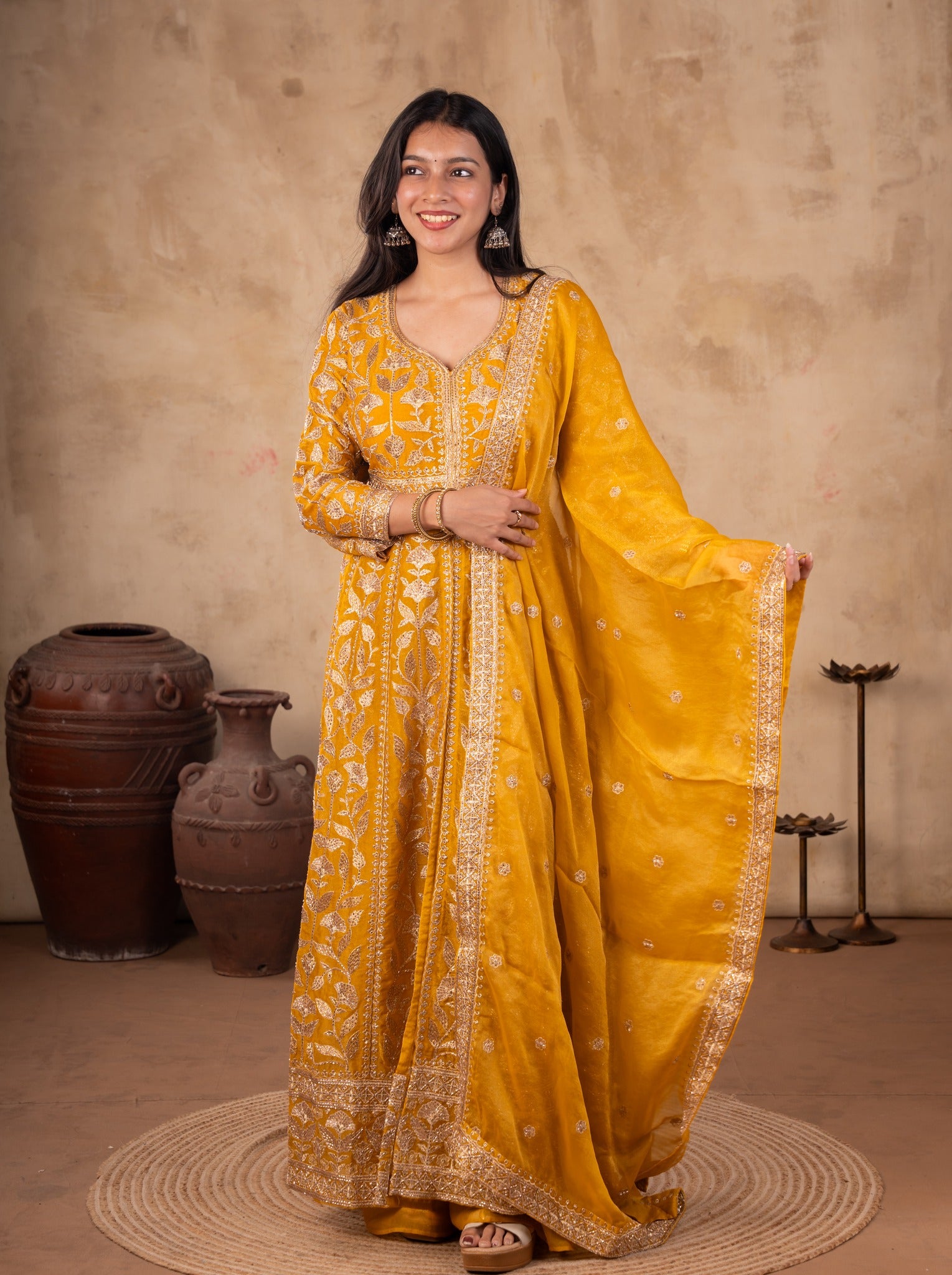 Front view of a model in a mustard tissue Anarkali suit with floral embroidery and a matching dupatta with an embellished border