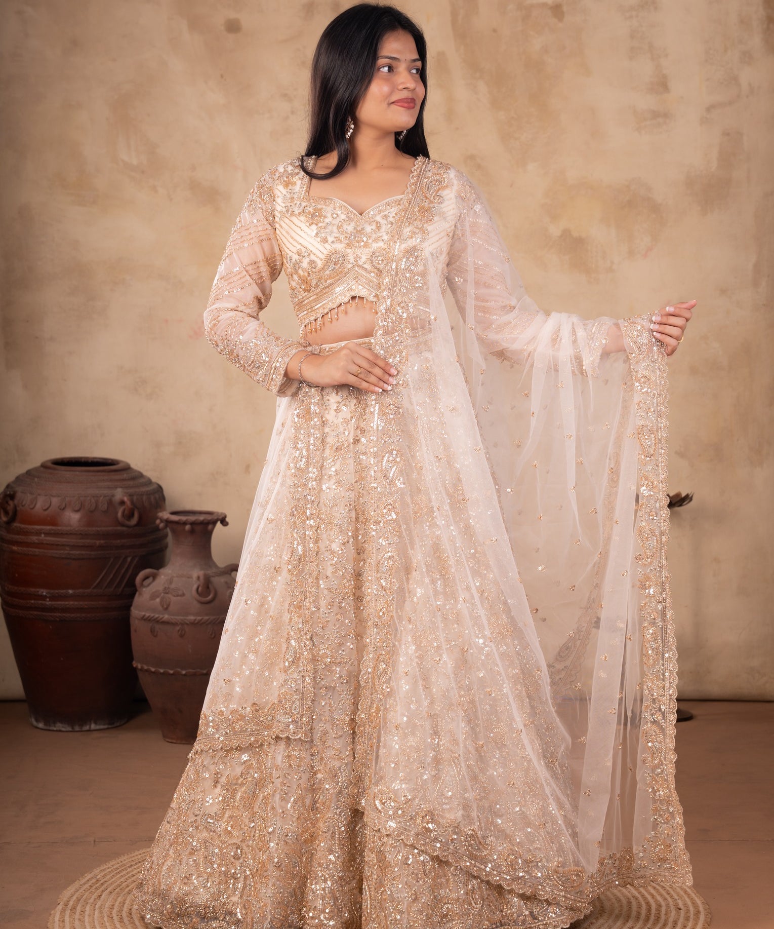 Model wearing a cream net lehenga with floral embellishments, a matching blouse with sheer sleeves, and a sheer dupatta with an embroidered border.