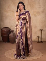 Model wearing a lustrous bronze-toned saree with a deep purple border, ornate gold floral and paisley embellishments, and a patterned pallu.