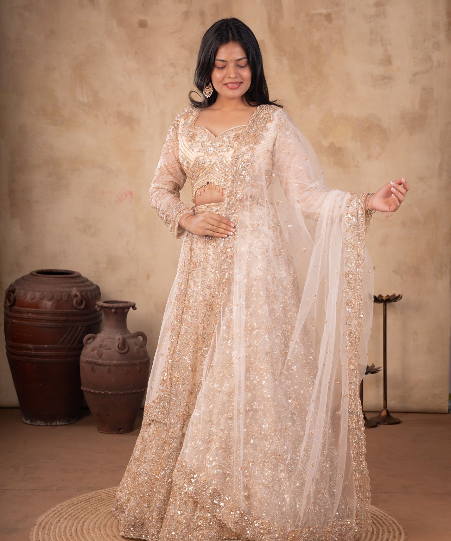 Model wearing a cream net lehenga with floral embellishments, a matching blouse with sheer sleeves, and a sheer dupatta with an embroidered border.