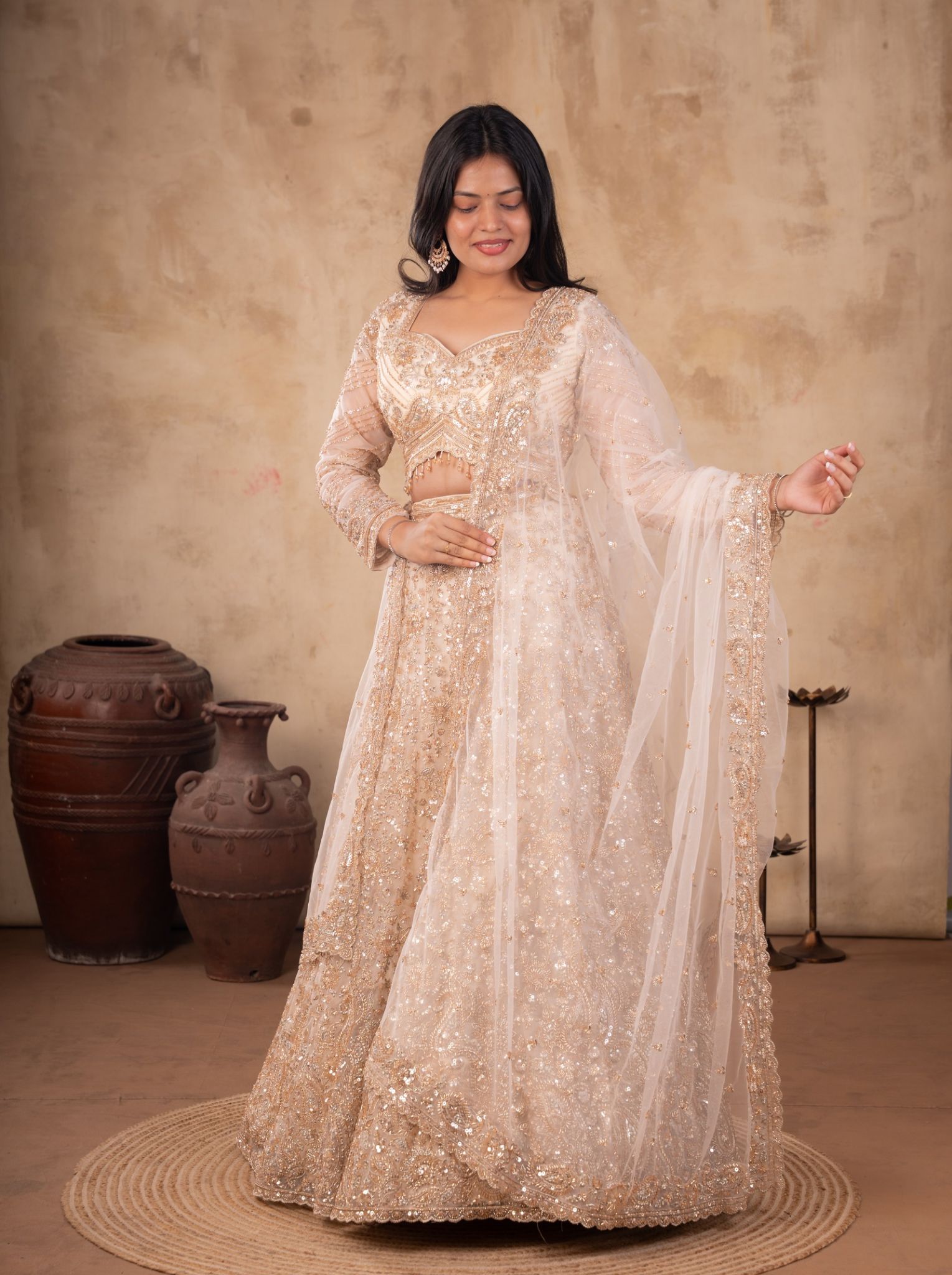 Model wearing a cream net lehenga with floral embellishments, a matching blouse with sheer sleeves, and a sheer dupatta with an embroidered border.