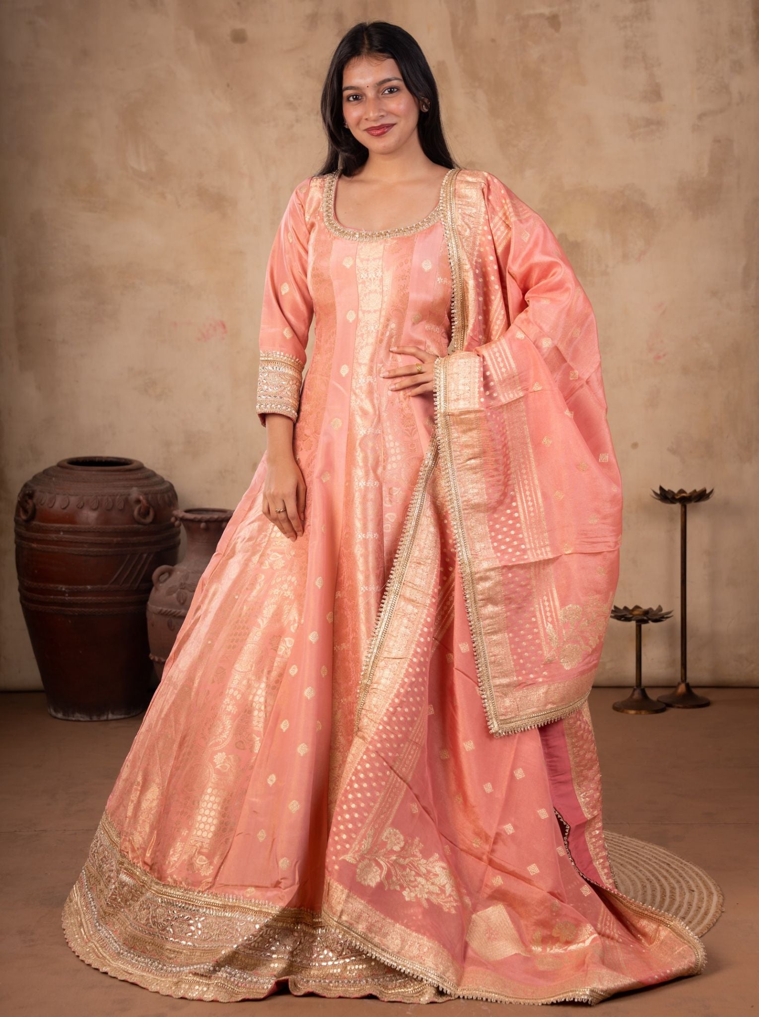 Model wearing a blush pink raw silk Anarkali with gold motifs, embellished neckline and cuffs, a wide gold hem, and matching bordered dupatta.
