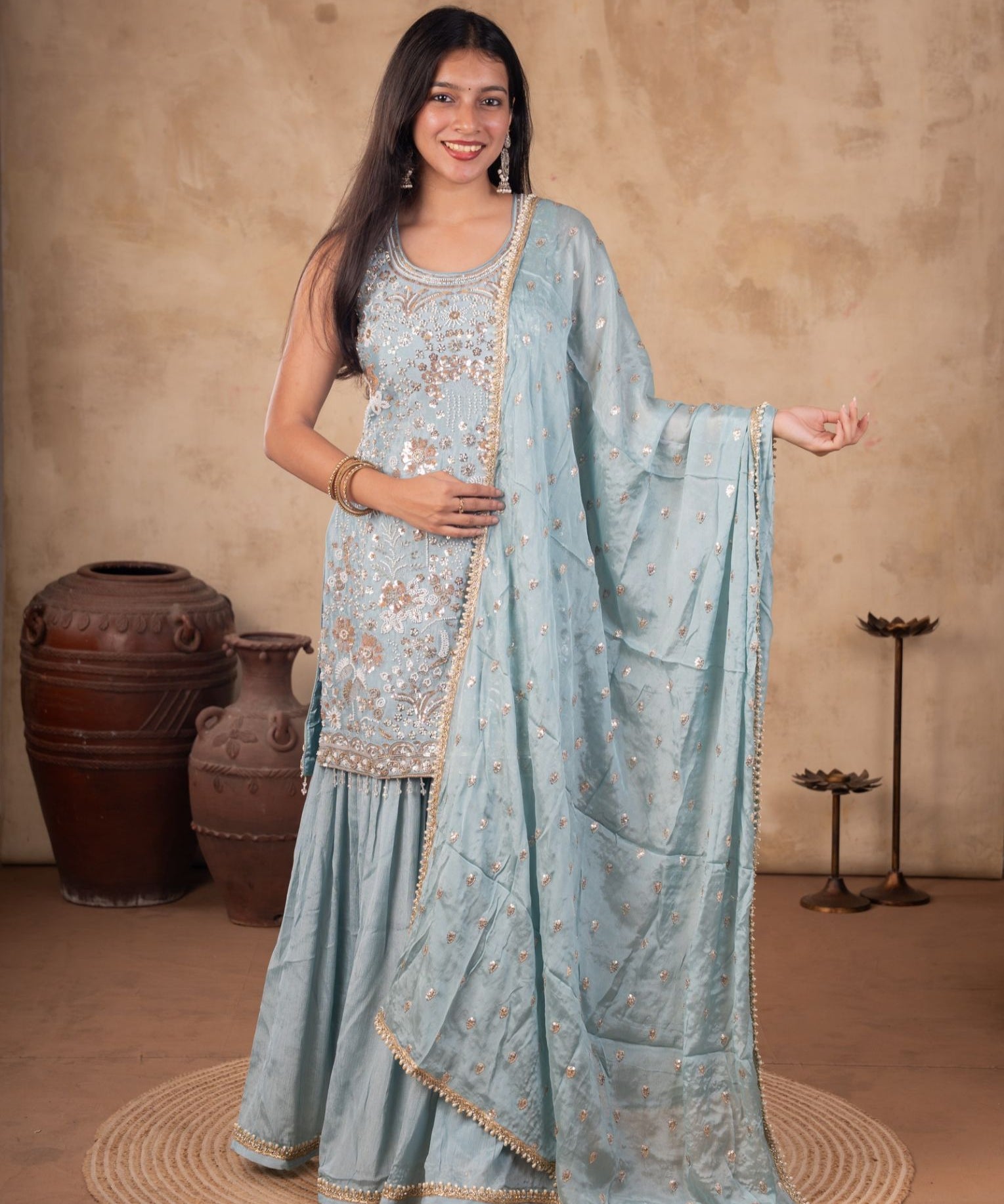 Model wearing a sage green georgette sharara suit with a sleeveless floral-embroidered kurta, flared trousers, and a matching dupatta with decorative trim.