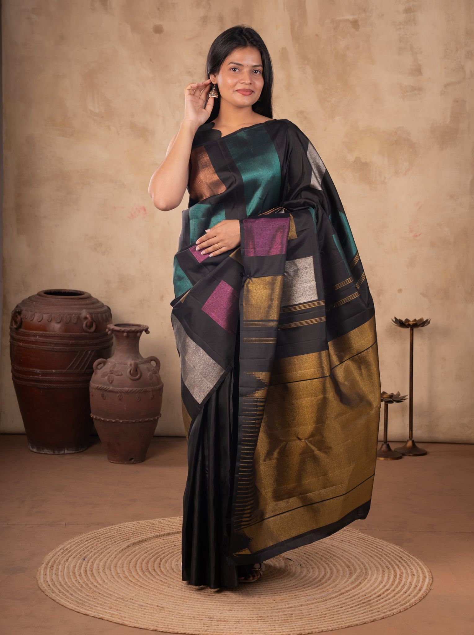 Model wearing a black soft silk saree with teal, purple and metallic square motifs, a broad gold pallu, and a black sleeveless blouse.