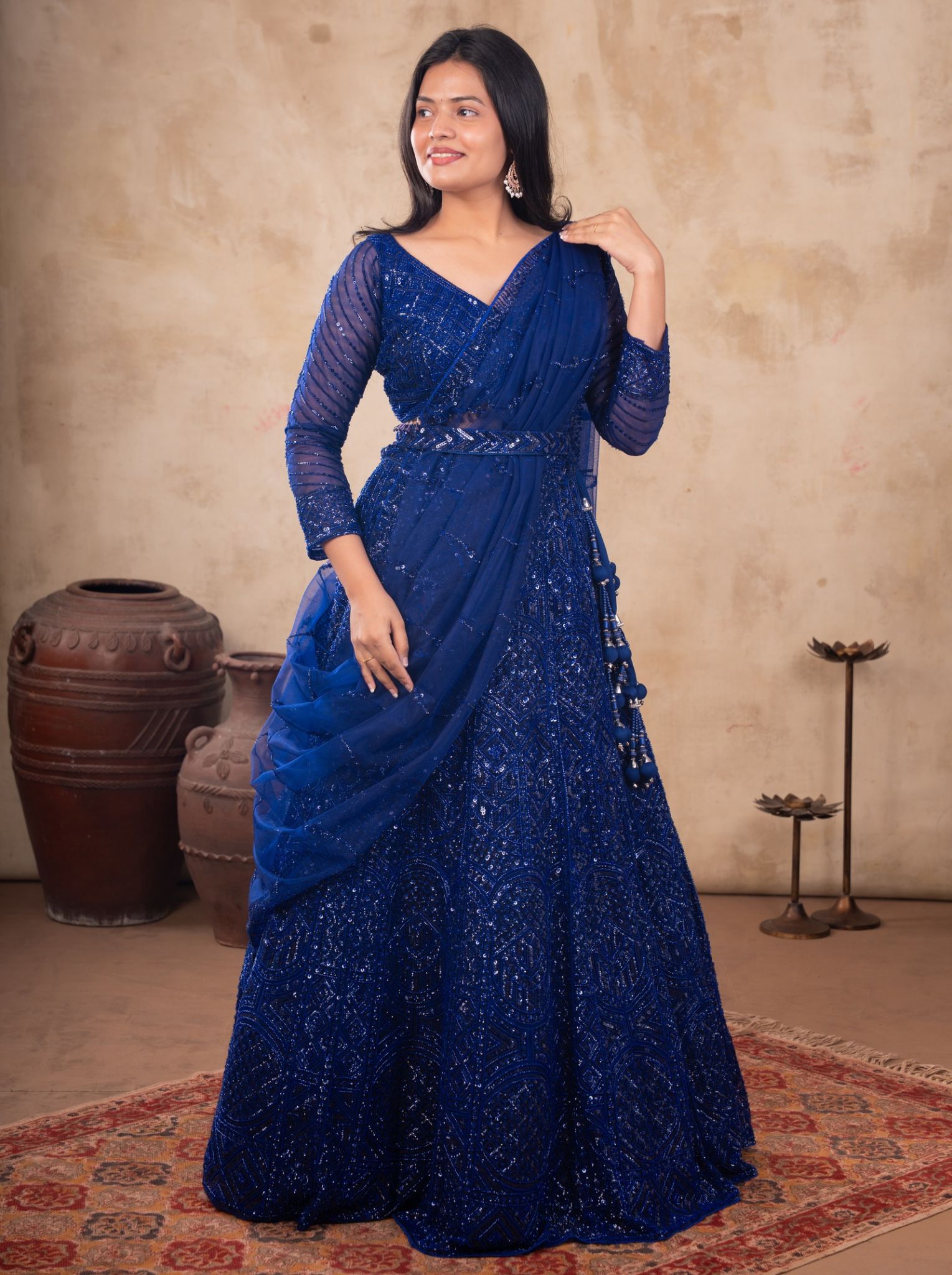 Model wearing a royal blue net lehenga with shimmering embellishments, a matching blouse with sheer sleeves, and a draped dupatta.