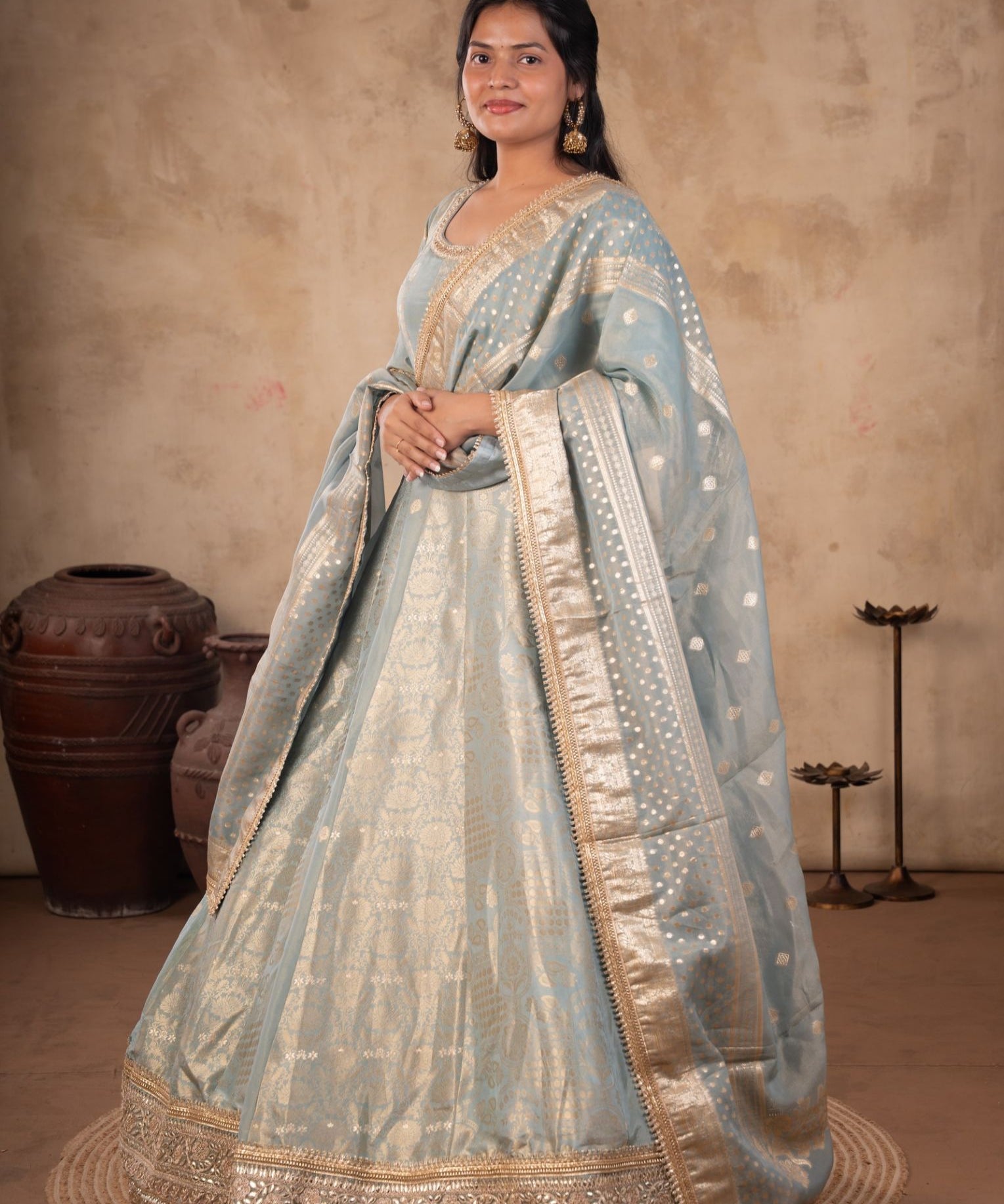 Model wearing a pale blue raw silk Anarkali with gold motifs, an ornate gold hem, and a matching dotted dupatta with a wide gold border.